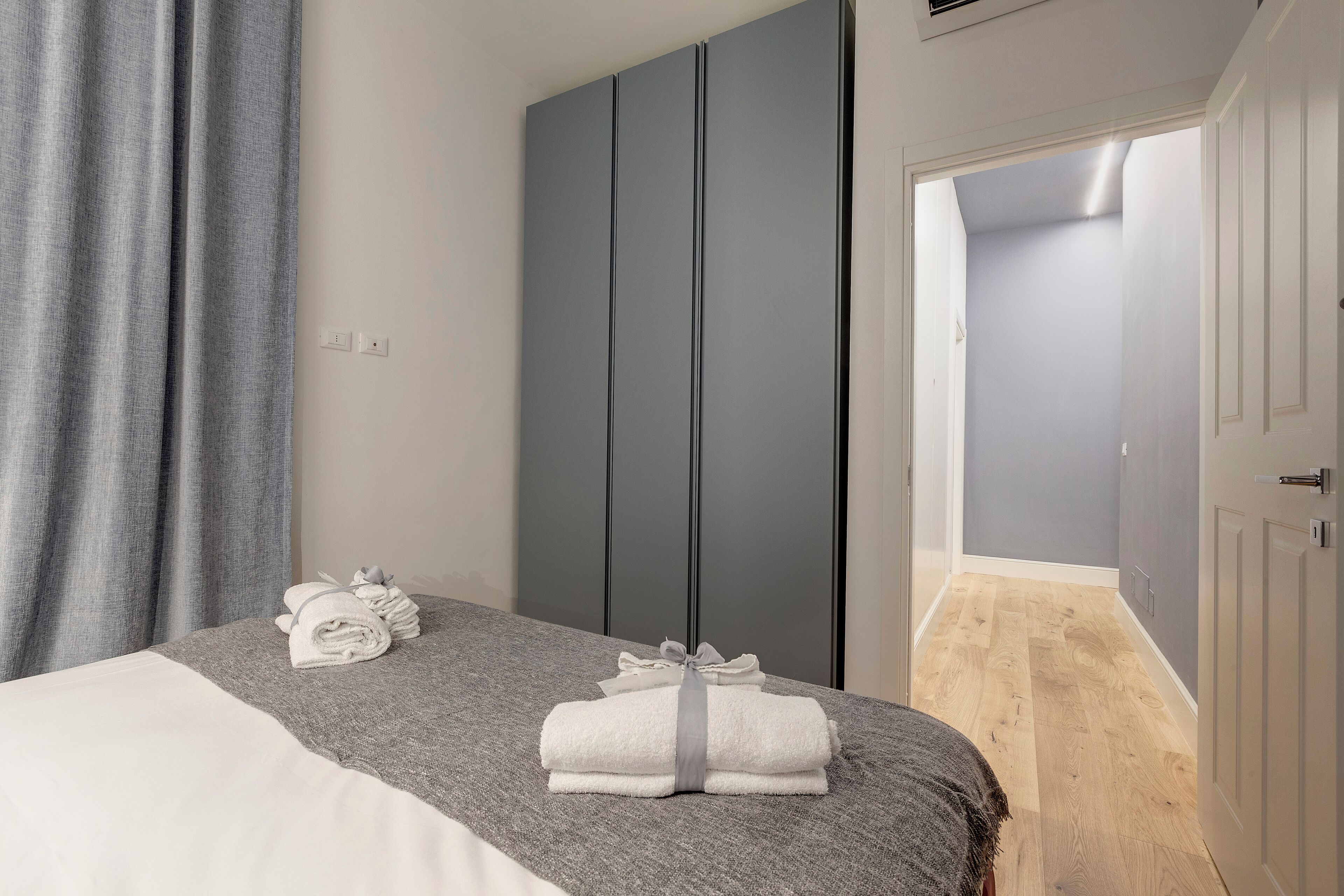 Un lit bien fait, avec du linge de lit blanc et gris, un rideau gris et une armoire grise, dans une chambre moderne dotée d’un plancher en bois et d’une porte donnant sur une autre pièce.