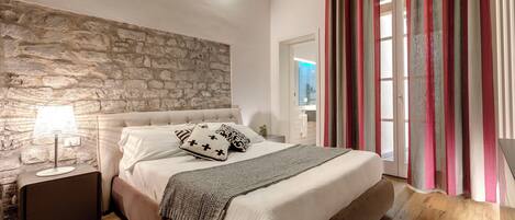 A bedroom with a stone wall, a bed with white bedding, a nightstand with a lamp, and a view of a bathroom through a glass door.