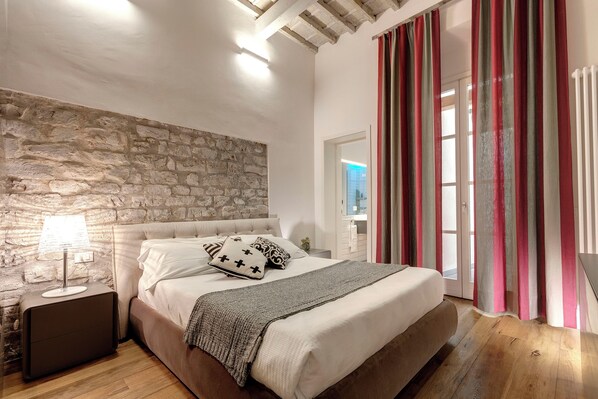 Un dormitorio con pared de piedra, cama con ropa blanca, mesita de noche con lámpara y vista a un baño a través de una puerta de vidrio.