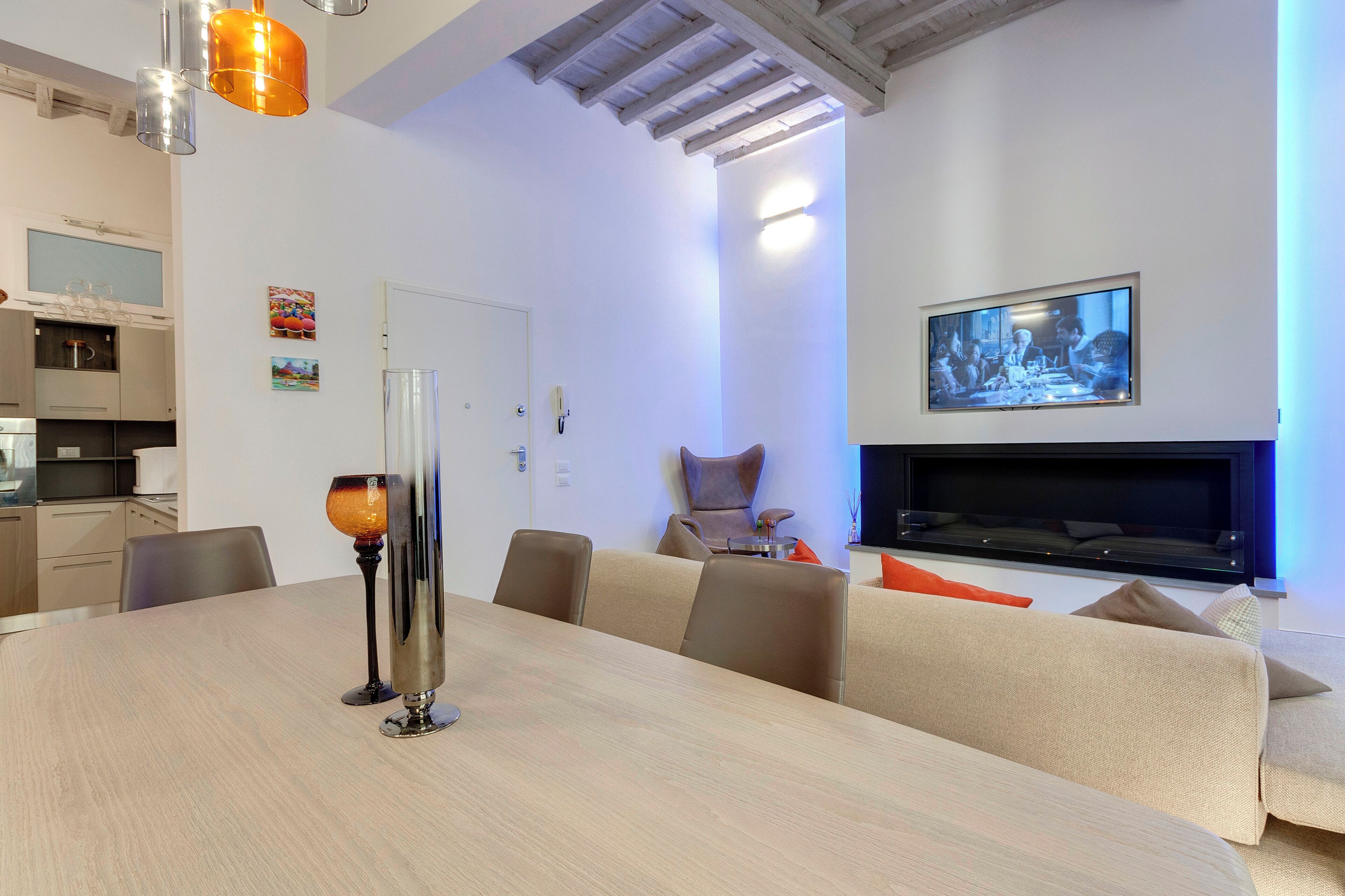 Un salon moderne doté d’une grande table à manger, de sièges confortables, d’un téléviseur à écran plat fixé au mur et d’un coin cuisine à l’arrière-plan.