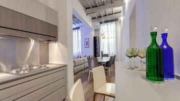 Una cocina moderna con extractor de acero inoxidable, un comedor con mesa y sillas, y dos botellas de vidrio coloridas sobre la mesa.