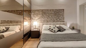 A bedroom with a stone wall, a bed with white bedding, a nightstand with a lamp, and a mirror on the wall. - Mamo Florence - Anita Apartment (Florence)