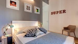 Een slaapkamer met een bed, kussens, een nachtkastje met een lamp, een stoel en ingelijste foto's aan de muur.
