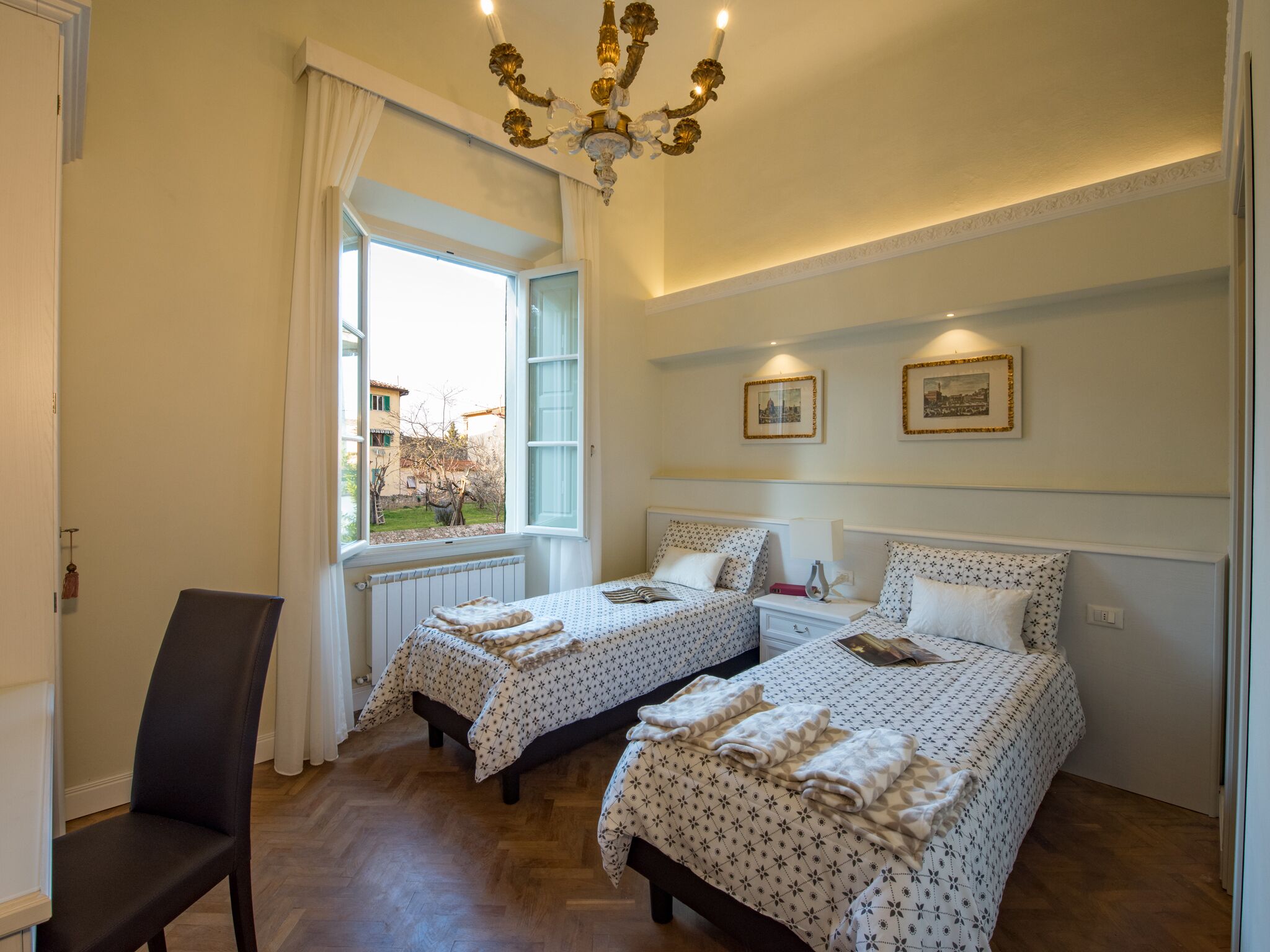 A bedroom with two beds, a chair, a window, and a chandelier.