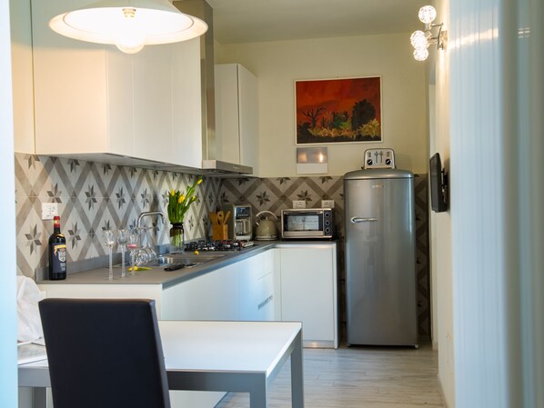 A compact kitchen with white cabinets, a microwave, a toaster, and a refrigerator. There is a dining table with chairs and a vase with flowers on the counter.