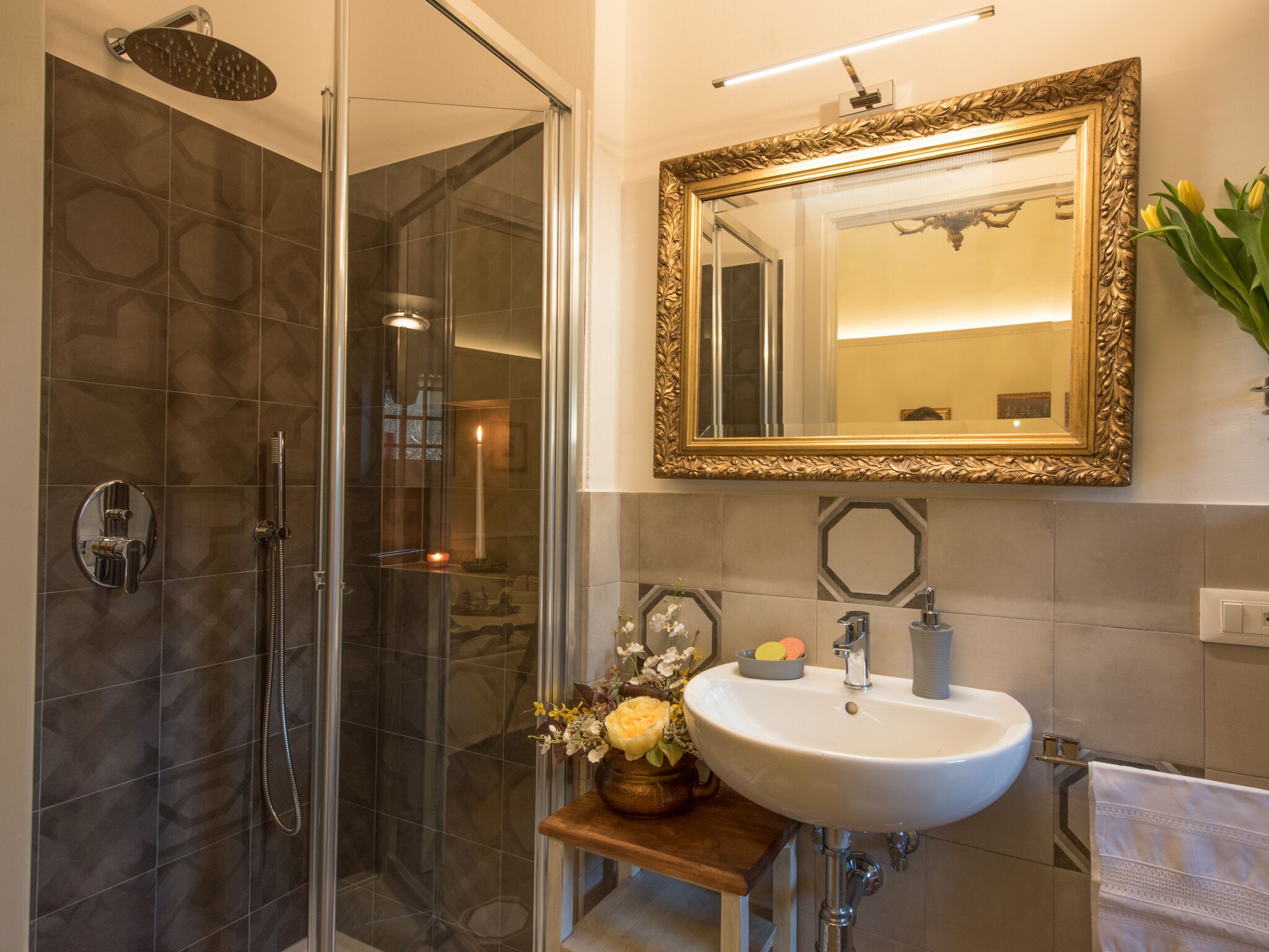 Een moderne badkamer met een douchecabine van glas, een witte wastafel met spiegel met gouden lijst, en een klein houten krukje met een bloemenvaas.