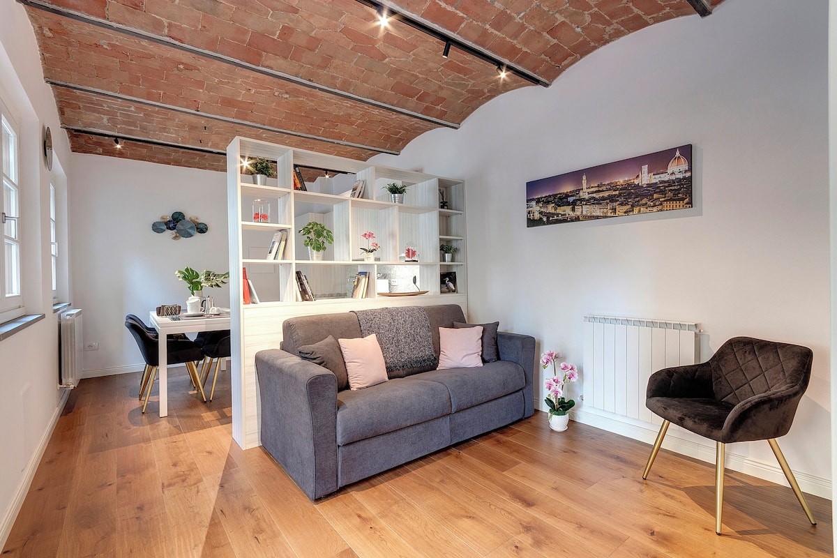 Una sala de estar moderna con un sofá gris, un sillón negro y una estantería blanca llena de plantas y libros.
