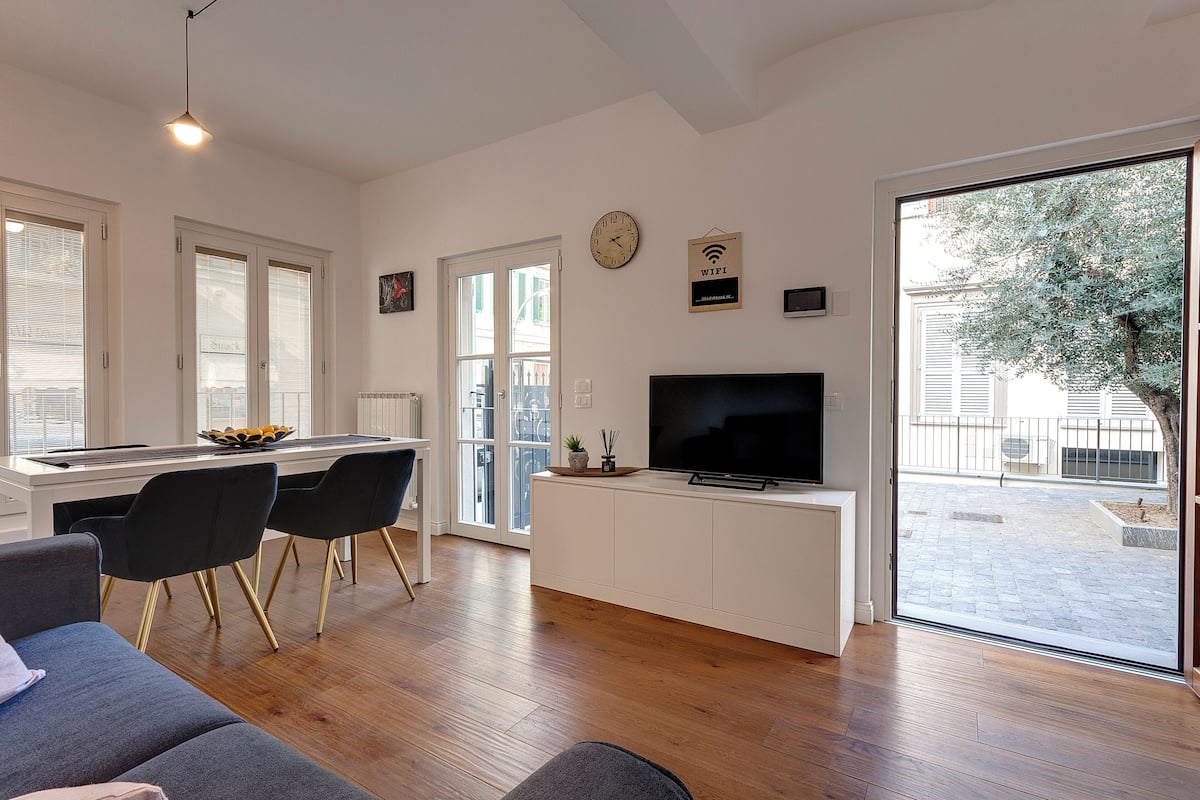 Una sala moderna con sofá, comedor con sillas, televisor en mueble bajo y un ventanal con vistas al exterior.