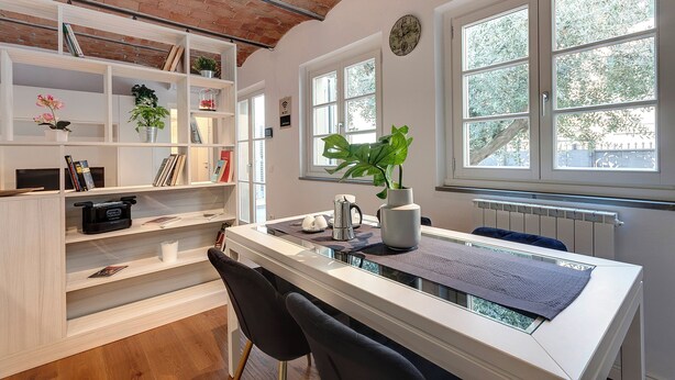 Una habitación moderna con un escritorio blanco, sillas negras, una estantería y un ventanal con vistas a árboles.