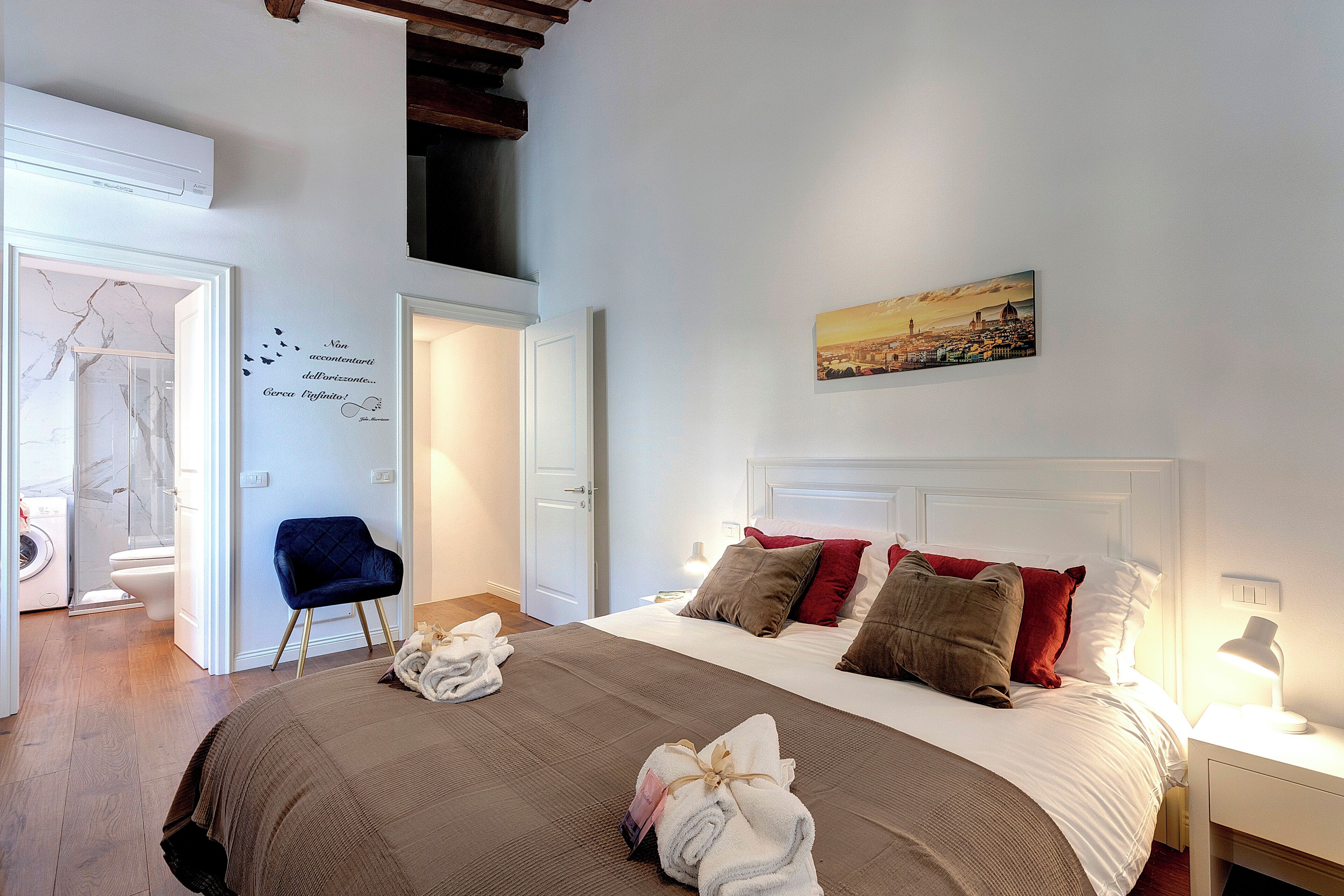 A hotel room with a large bed, a cityscape painting, a wall decoration with a quote, and a blue chair.