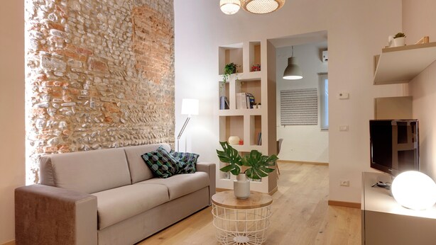 Een moderne woonkamer met een stenen accentmuur, een beige bank, een ronde salontafel met planten en een televisie op een dressoir.