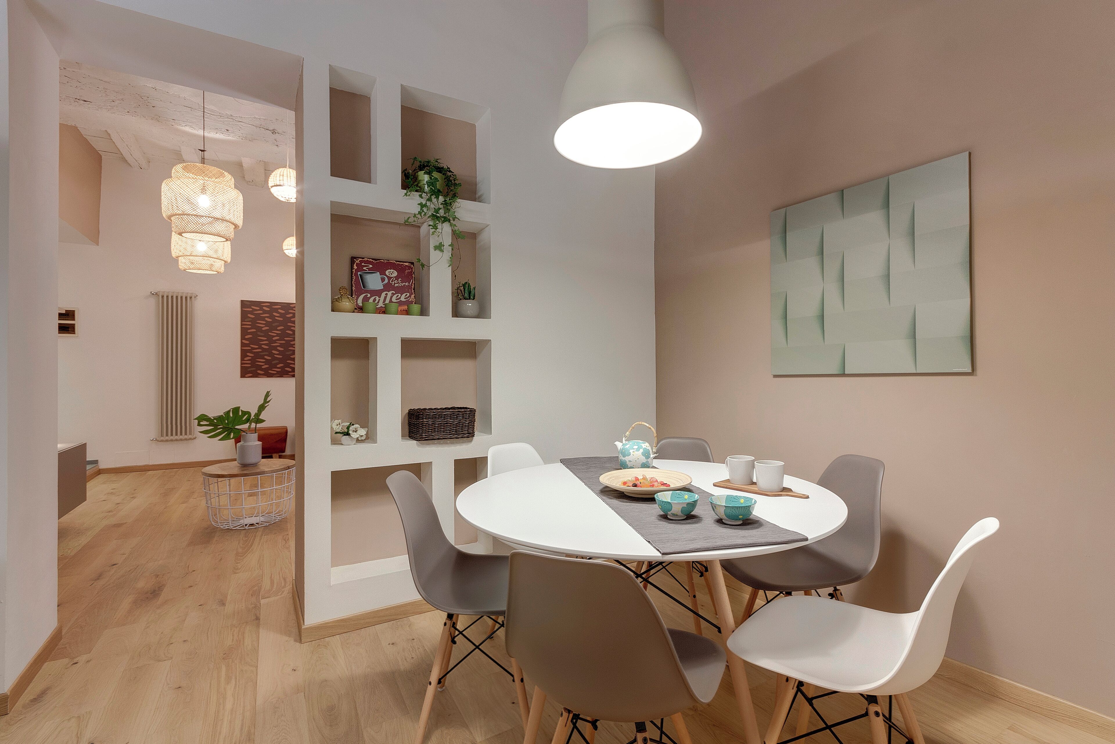 A dining area with a round table, four chairs, a hanging light fixture, and a wall-mounted shelf with decorative items.