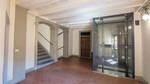 Ένας διάδρομος με πλακάκια στο δάπεδο, μια σκάλα με μεταλλικό κάγκελο και ένας ανελκυστήρας με γυάλινο θάλαμο.