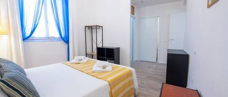 A hotel room with a bed, a bedside table, a chair, a wardrobe, a desk, and a door leading to another room.