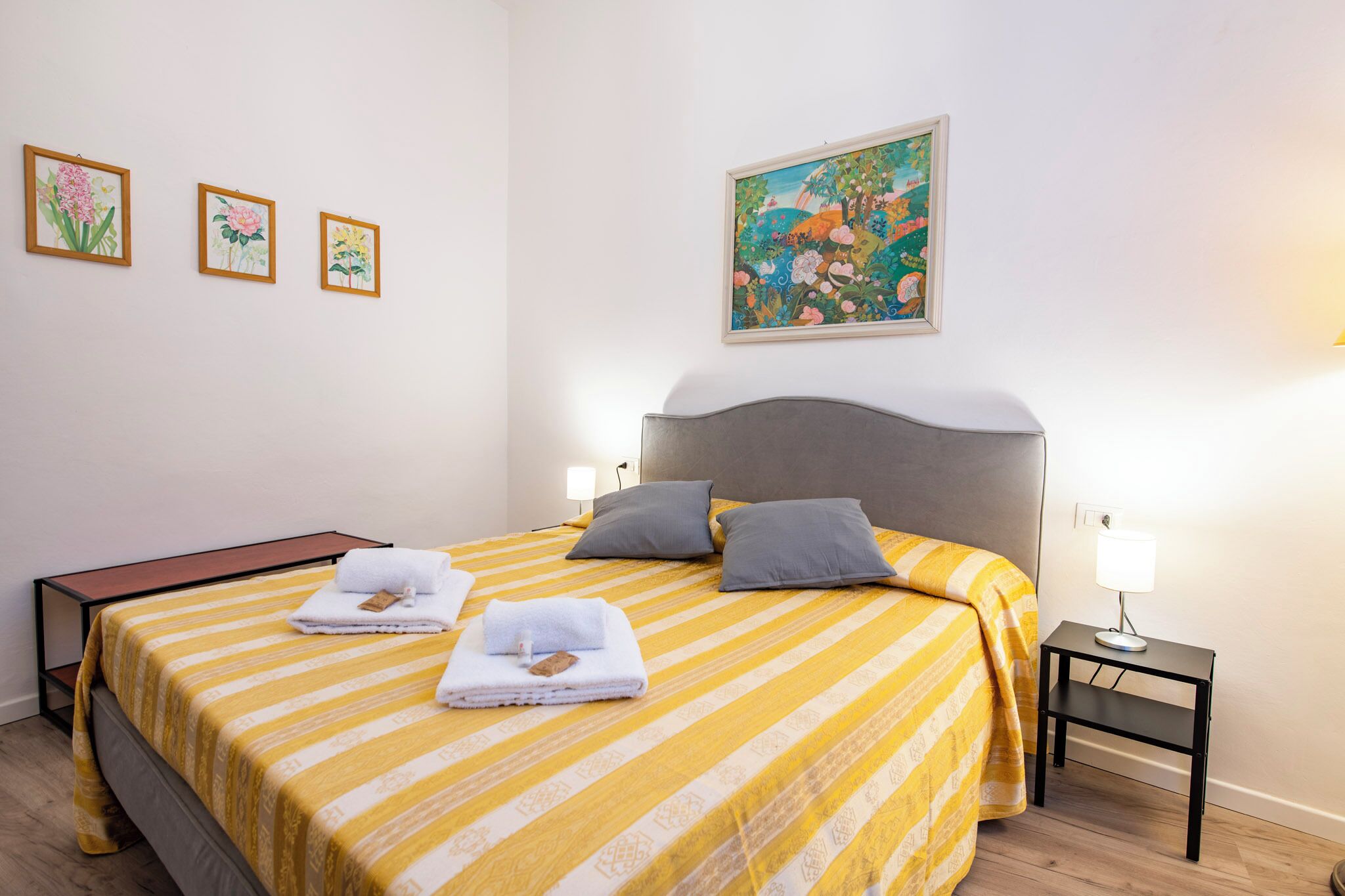 A hotel room with a bed, pillows, a bedside table with a lamp, and three framed floral artworks on the wall.