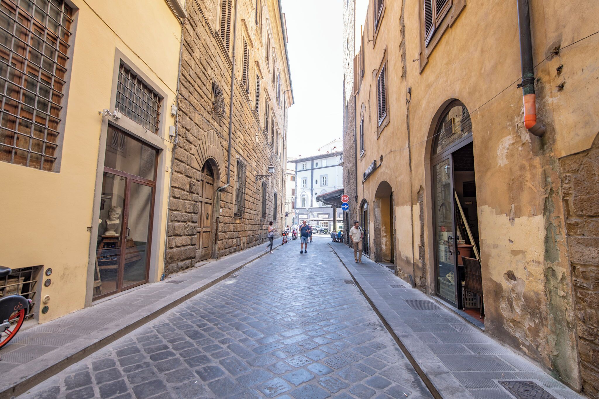A narrow cobblestone street flanked by historic buildings with arched doorways and large windows.