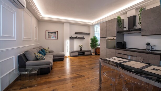 A modern living room with a grey sofa, wooden flooring, and a dining area with a glass table and chairs. - Mamo Florence - Cherubini Apartment (Florence)