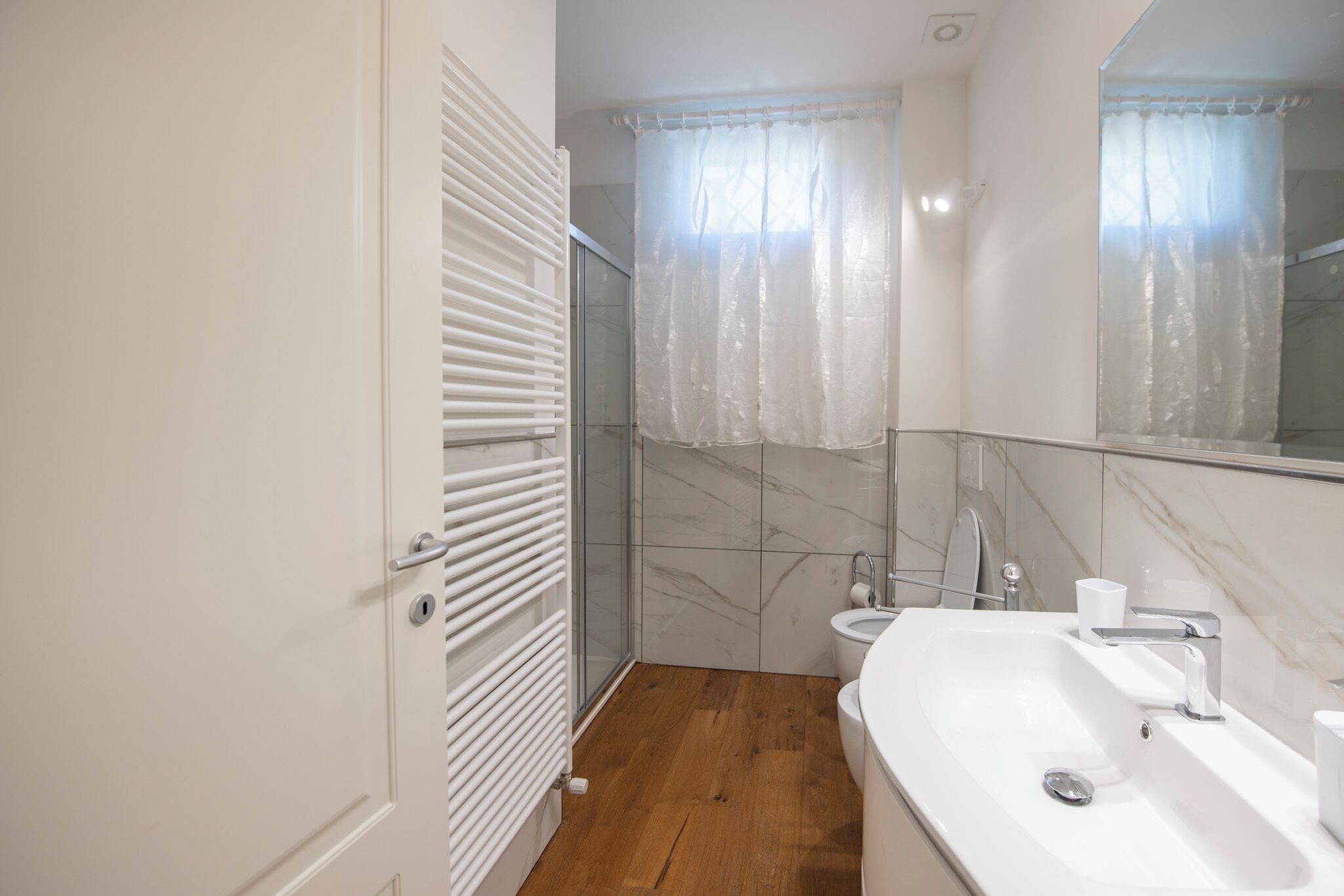 A bathroom with a wooden floor, white fixtures, a glass shower enclosure, and a large mirror.