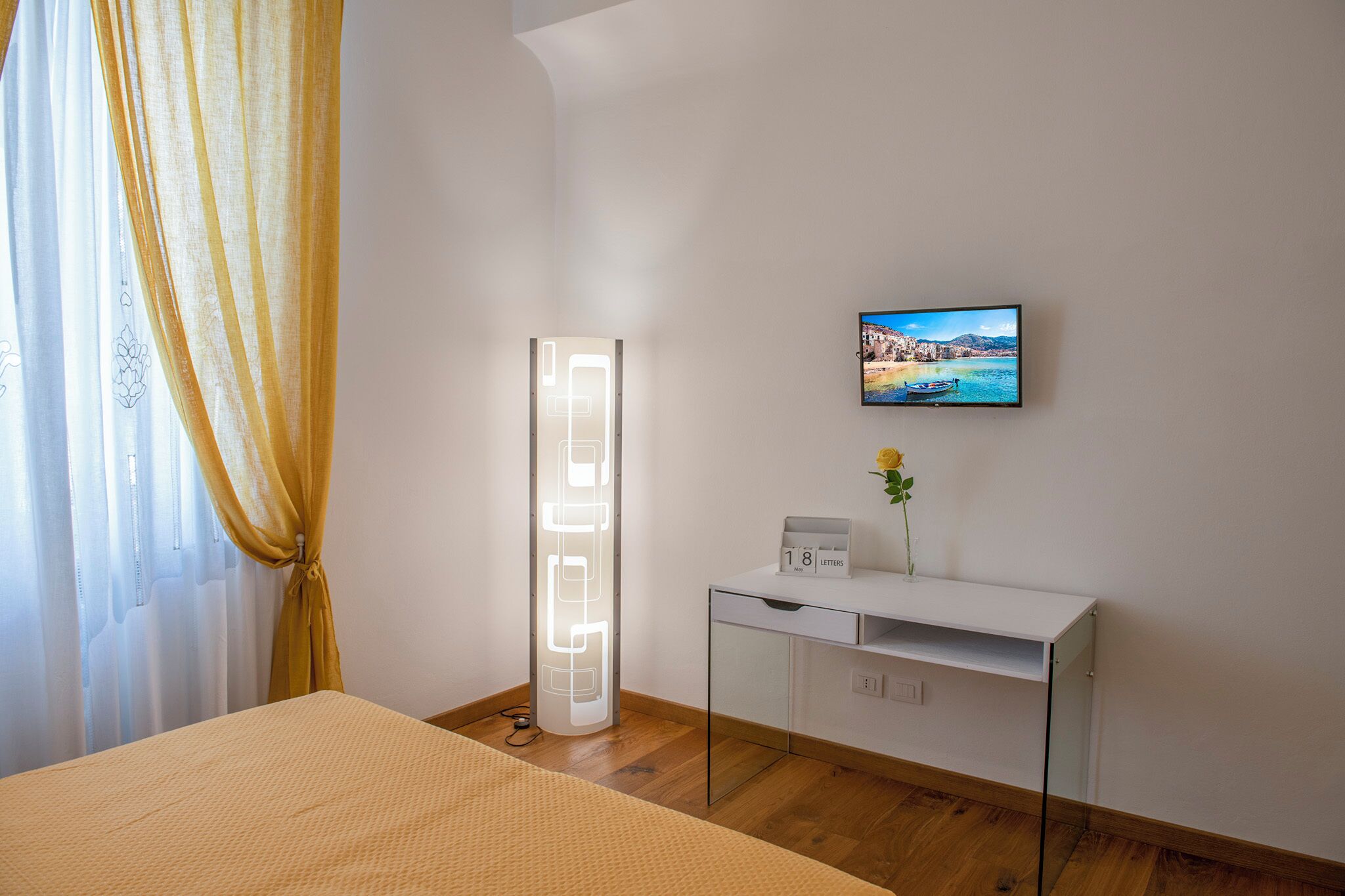 A modern hotel room with a bed, a bedside table with a clock, a picture on the wall, and a floor lamp.