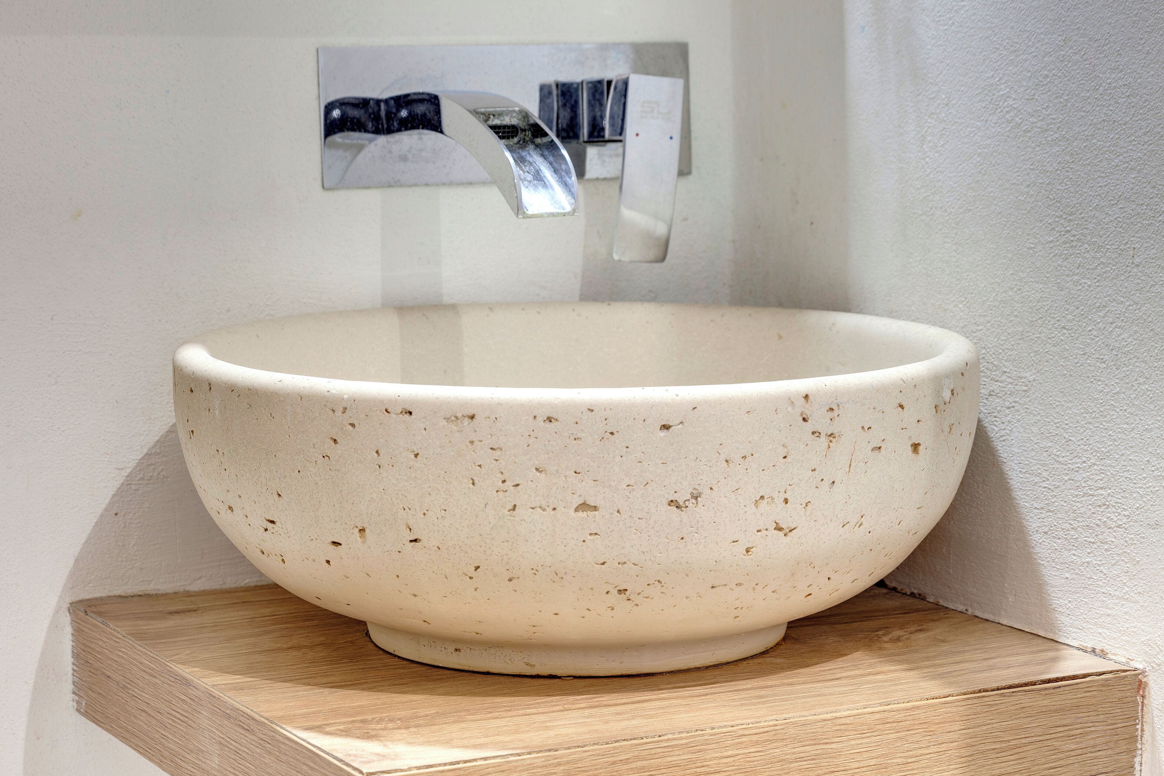 A white ceramic bowl with speckles, placed on a wooden shelf against a wall with a metal faucet and a small picture frame.