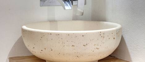 A white ceramic bowl with speckles, placed on a wooden shelf against a wall with a metal faucet and a small picture frame.