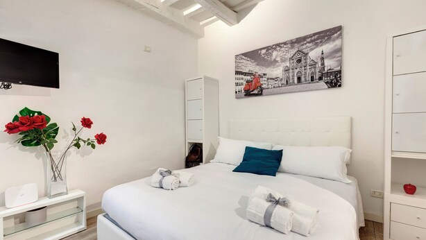 A neatly made bed with white linens and two pillows, a bedside table with a vase of red flowers, a wall-mounted TV, and a painting of a cityscape. - Mamo Florence - Cristofano Studio (Florence)