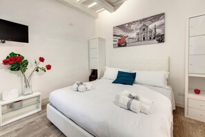 A neatly made bed with white linens and two pillows, a bedside table with a vase of red flowers, a wall-mounted TV, and a painting of a cityscape. - Mamo Florence - Cristofano Studio (Florence)