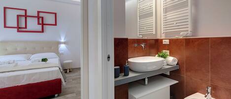 Une chambre moderne avec un lit, une table de chevet et un mur d’accent rouge. La salle de bain est équipée d’un grand lavabo, d’un miroir et d’un porte-serviettes.