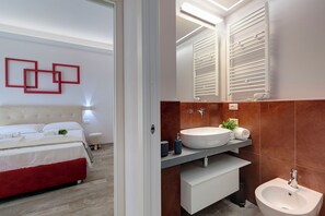 A modern bedroom with a bed, bedside table, and a red accent wall. The bathroom features a large sink, mirror, and towel rack. - Mamo Florence - Dante & Virgilio Apartments (Florence)