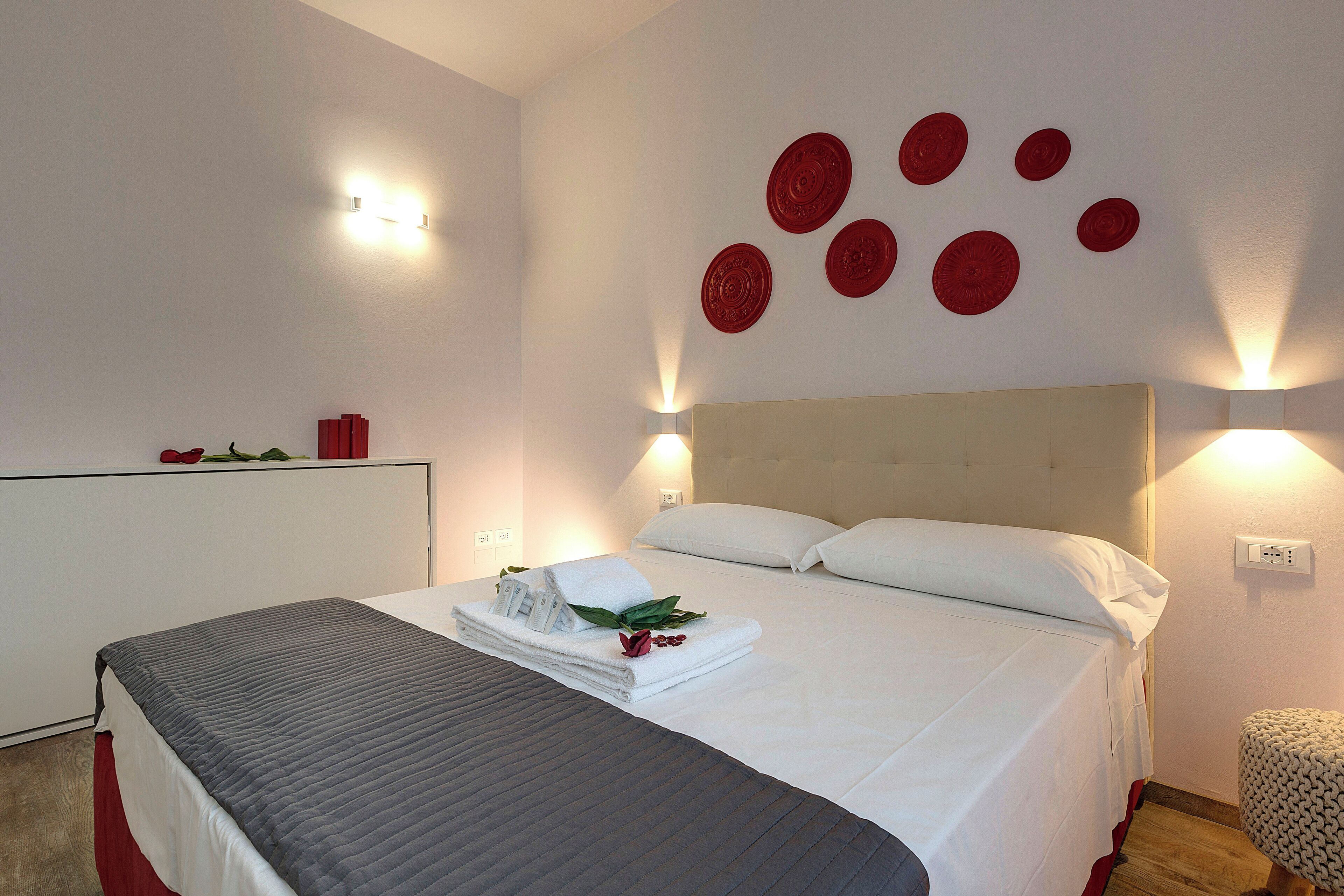 Uma cama bem arrumada com lençóis brancos e um cobertor cinzento, uma mesa de cabeceira com uma luminária, e uma parede com detalhes circulares.