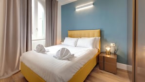 Una cama bien hecha con sábanas blancas y dos toallas, una mesita de noche con una lámpara y una ventana con cortinas.