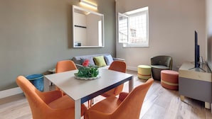 Una sala moderna con un sofá, sillas naranjas, una mesa con un centro de flores, un espejo y un televisor.
