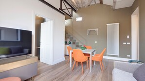 Una sala moderna con un televisor de pantalla plana, un sofá, un comedor con sillas naranja y una escalera que sube a otro nivel.
