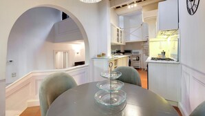 Un área de comedor con un pastelero de vidrio sobre una mesa redonda, una araña y una cocina al fondo.