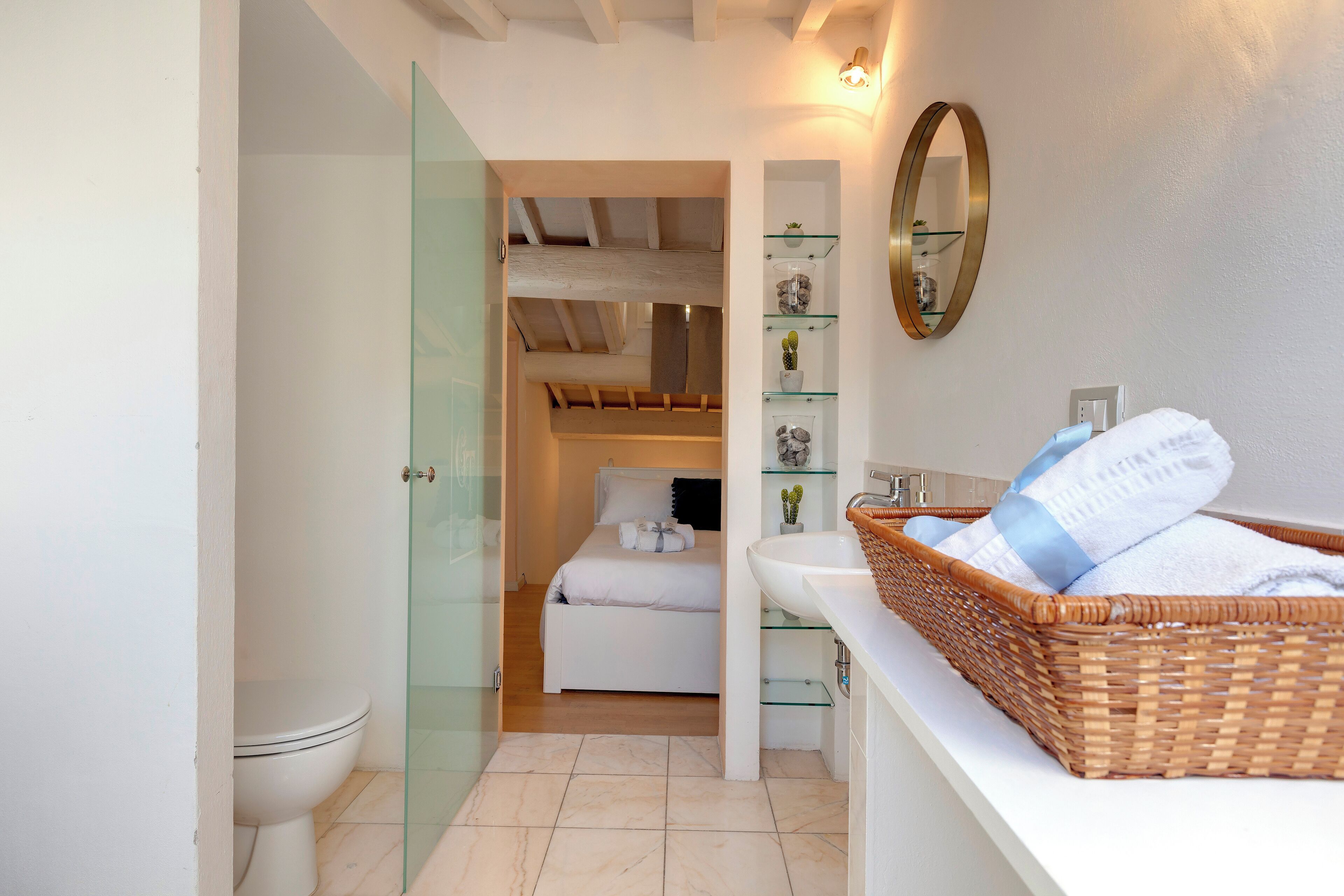 A modern bathroom with a glass shower enclosure, a white toilet, a glass-walled walk-in closet, a bed with white bedding, and a wicker basket on the vanity.