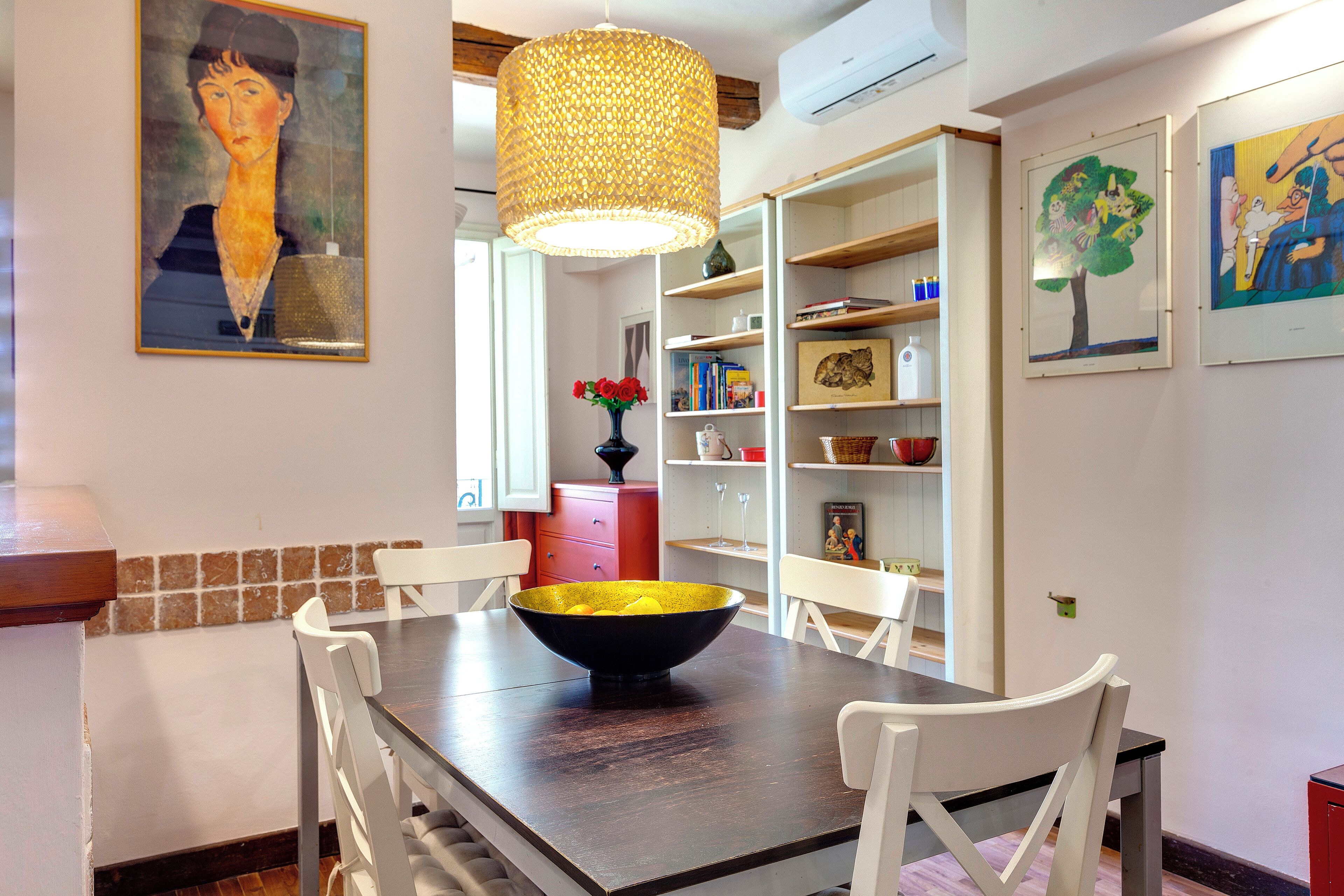 Uma área de refeições com uma mesa de madeira escura, cadeiras brancas, uma tigela preta grande e um lustre tecido pendurado.