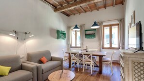 Ein Wohnzimmer mit einer Couch, einem runden Holztisch und einem Esstisch mit Stühlen. Ein Fernseher steht auf einer Kommode und es gibt ein Fenster mit Vorhängen.