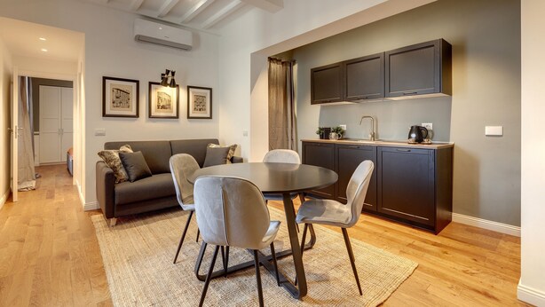 A modern living room with a round dining table, grey sofas, and a dark kitchen with built-in cabinets. - Mamo Florence - La Badessa Apartment (Florence)