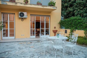 An outdoor patio with white metal chairs and a table, a yellow building with a door and windows, and potted plants on the balcony. - Mamo Florence - La Galerie Luxury Apartment (Florence)