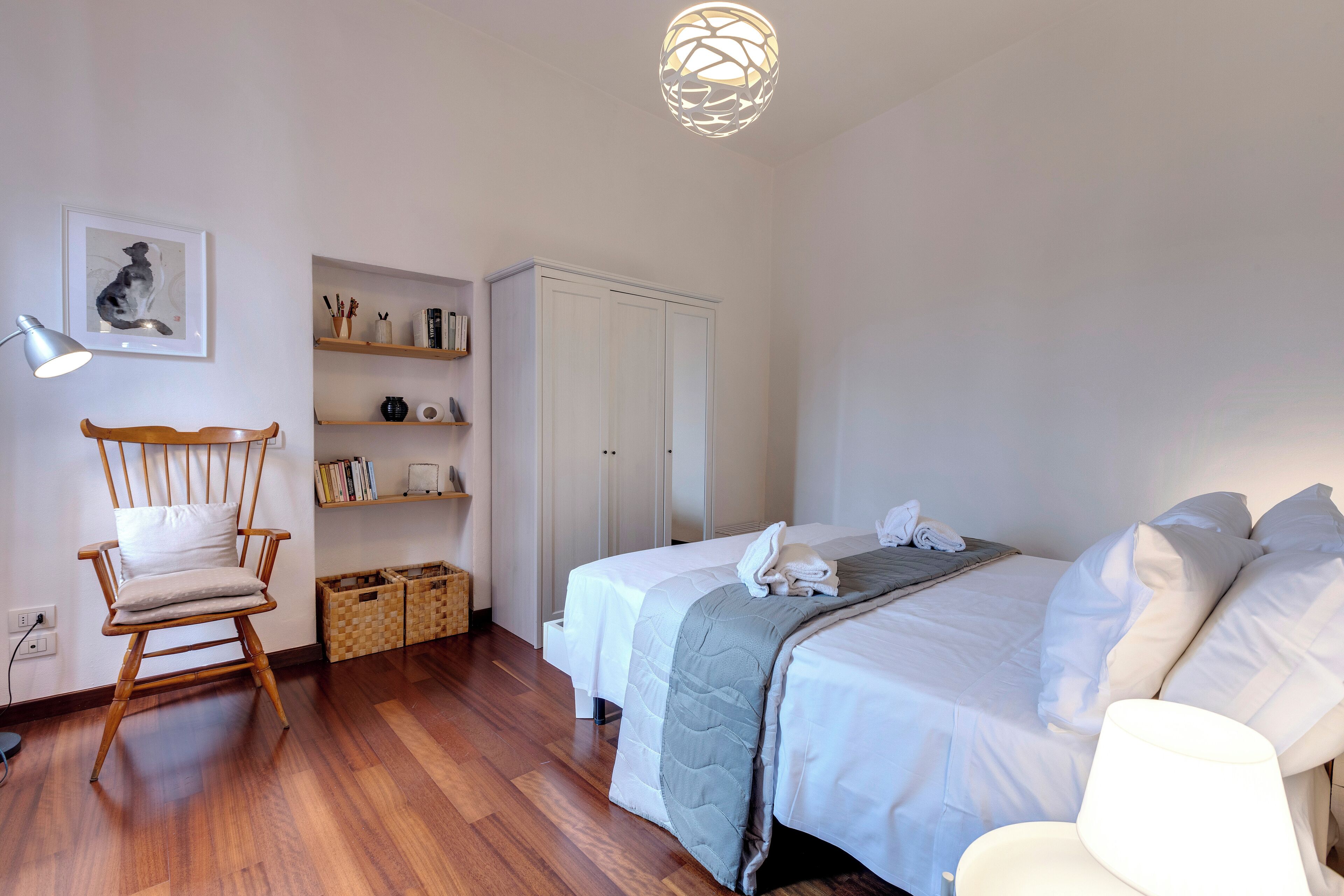 A bedroom with a bed, a chair, a desk, a lamp, and a shelf with books and decorative items.