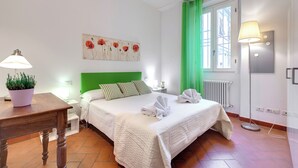 A neatly made bed with a green headboard, a bedside table with a lamp, a potted plant, and a painting on the wall. - Mamo Florence - Maruffi Apartment (Florence)
