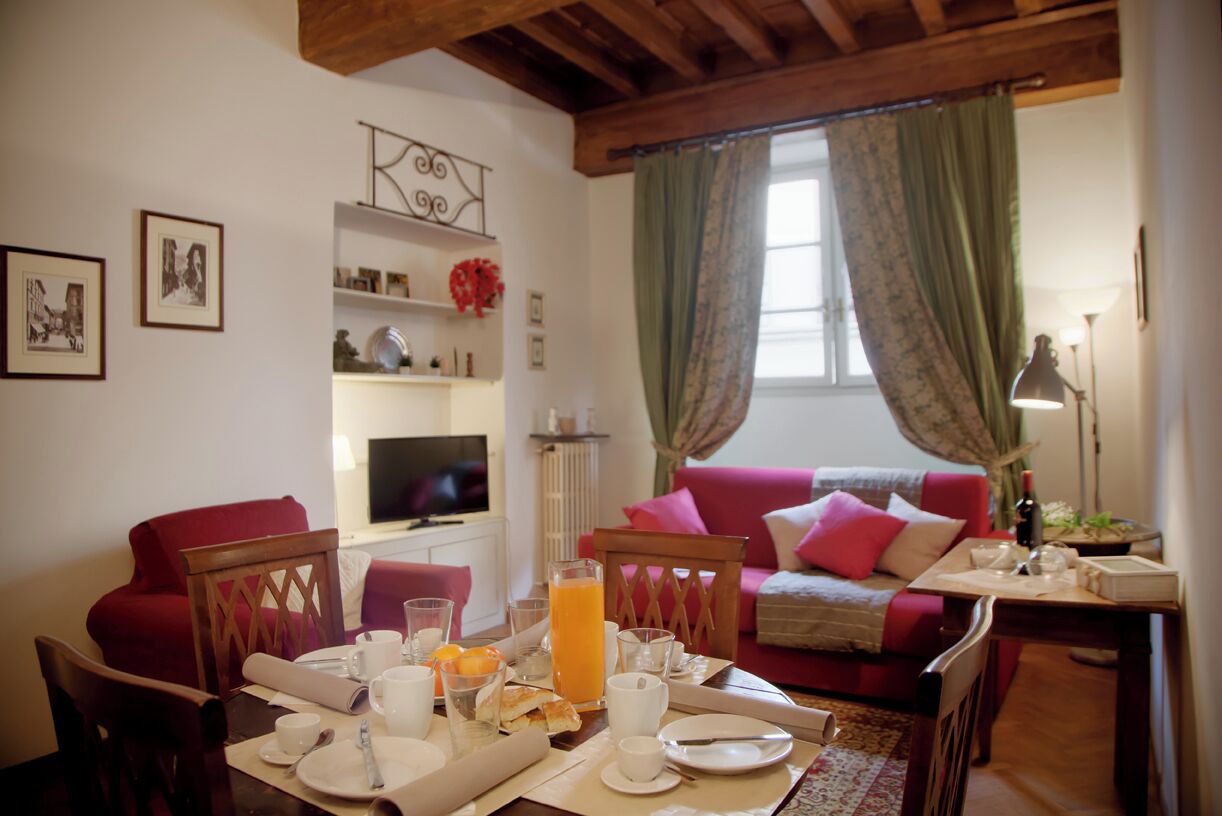 Un comedor con una mesa dispuesta para el desayuno, un televisor, un sofá y una ventana con cortinas.