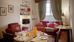 A dining area with a table set for breakfast, a television, a sofa, and a window with curtains. - Mamo Florence - Palazzo Spinelli Apartment (Florence)