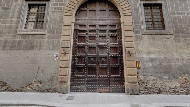 A large, arched wooden door with metal reinforcements, flanked by two smaller windows with metal bars, set in a stone building with a plain facade. - Mamo Florence - Palazzo Spinelli Apartment (Florence)