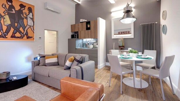A modern living room with a grey sofa, a brown ottoman, and a dining area with a white table and chairs. - Mamo Florence - Parione Apartment (Florence)