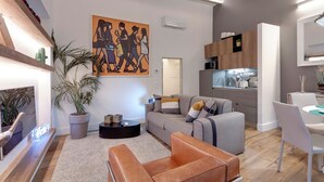 A modern living room with a grey sofa, a brown ottoman, a coffee table, and a large painting on the wall. There is a kitchen area with wooden cabinets and a dining table with chairs. - Mamo Florence - Parione Apartment (Florence)