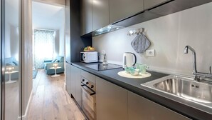 Una cucina moderna con lavello in acciaio inossidabile, forno a microonde e bollitore sul piano di lavoro. Si vede una finestra con vista su un soggiorno sullo sfondo.