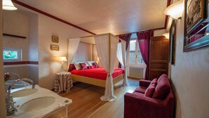 Une chambre à coucher avec un lit à baldaquin, un coin salon avec un canapé rouge, une grande fenêtre avec des rideaux et une salle de bain avec un lavabo et un miroir.