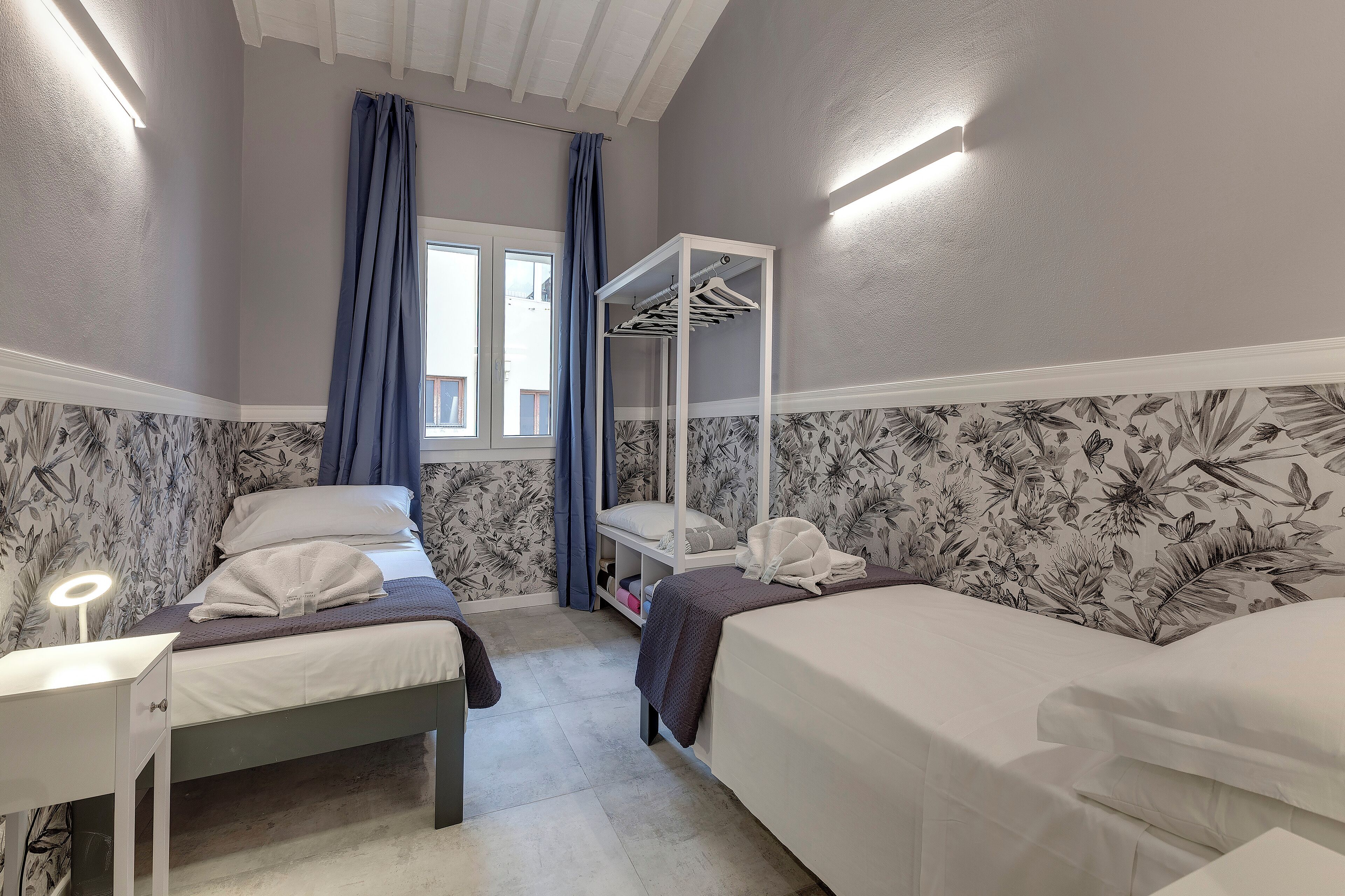 A hotel room with two beds, a wardrobe, a vanity with a mirror, and a window with curtains.