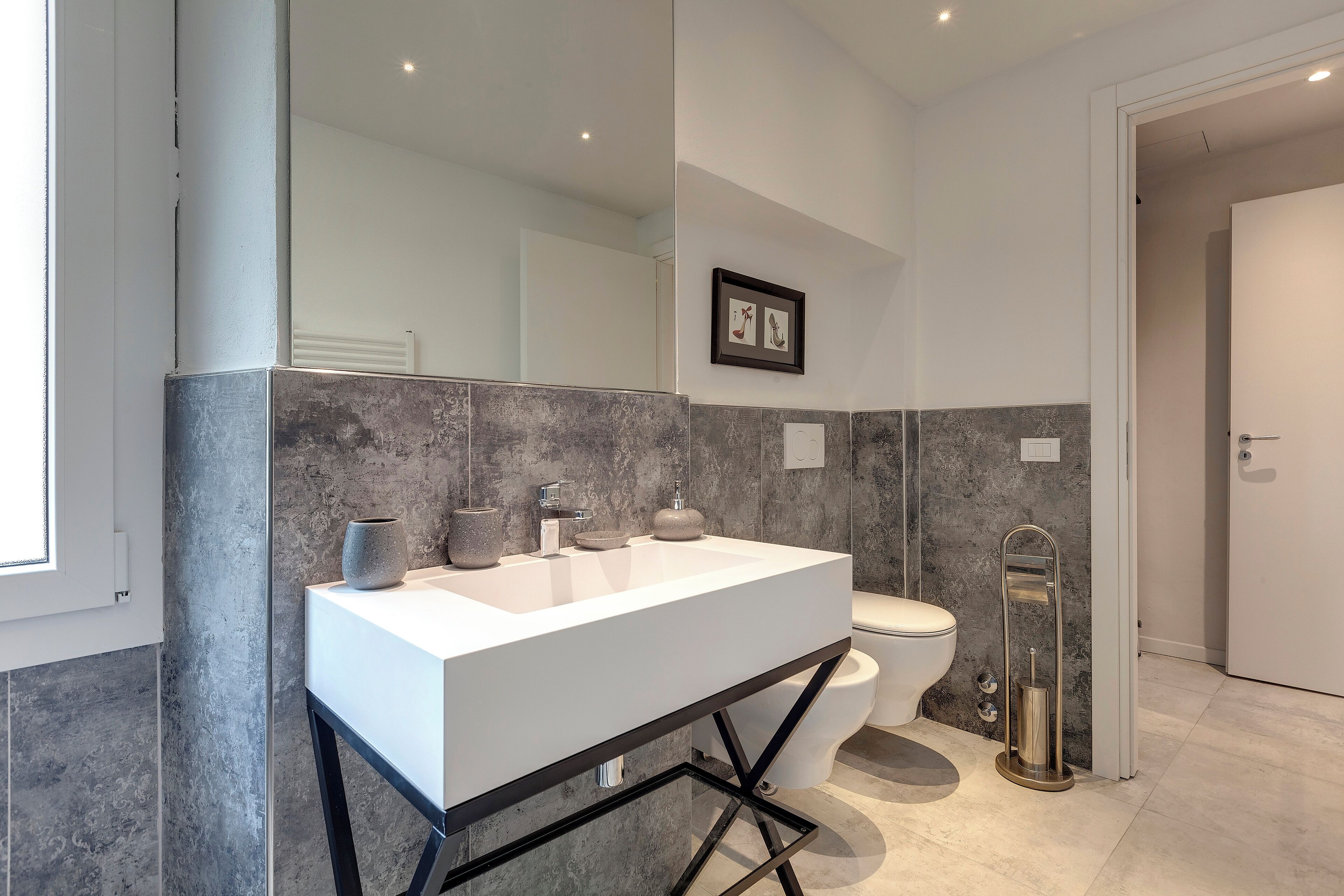 A modern bathroom with a white sink, dark tiled walls, a toilet, and a glass-encased mirror.